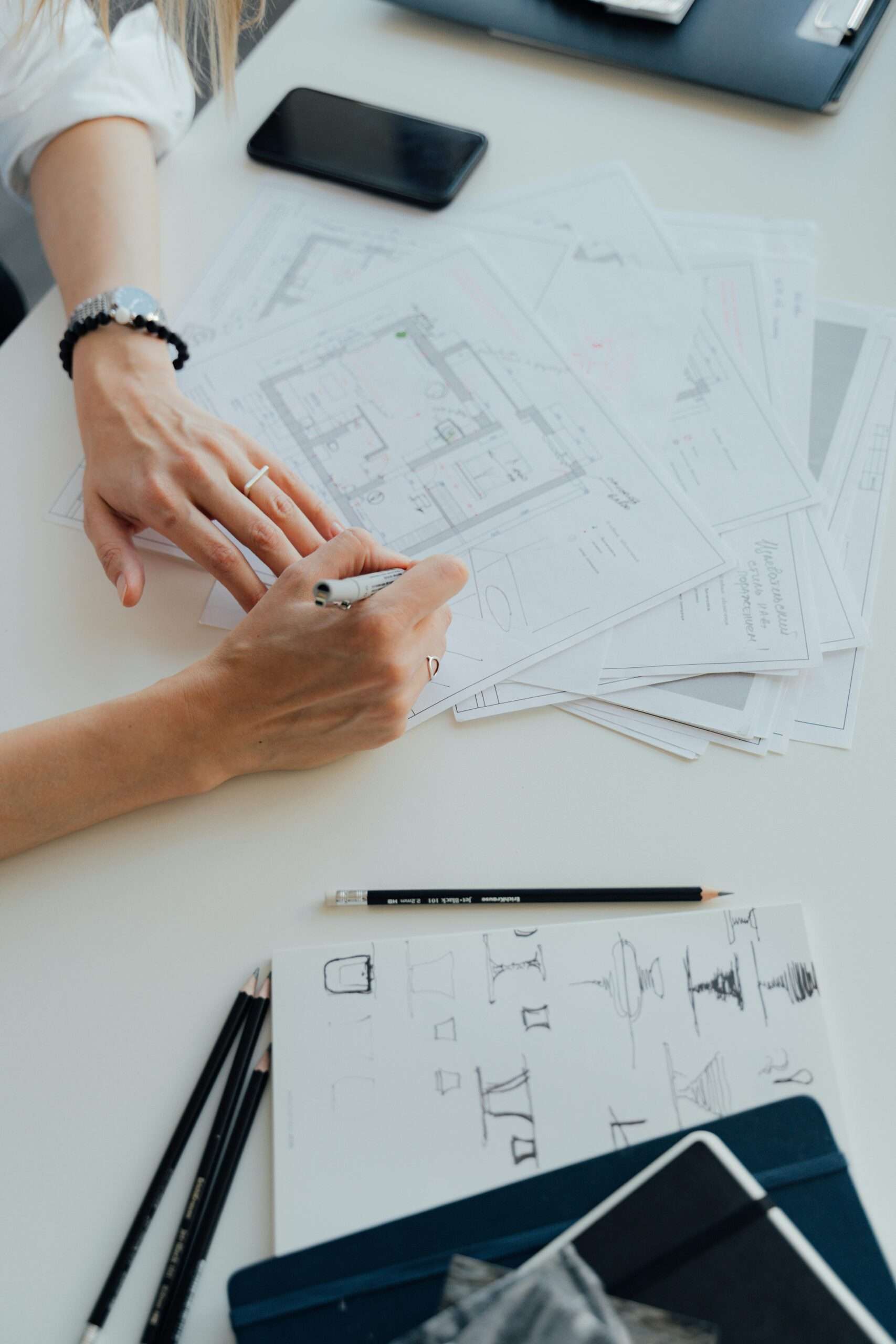 Des mains dessinent des plans d'architecture sur un bureau avec des papiers et des plans éparpillés. Des crayons et un carnet sont à proximité, à côté d'un smartphone. La personne tient un stylo, se concentrant sur des dessins détaillés au milieu de l'espace de travail organisé, suggérant un environnement animé et créatif.