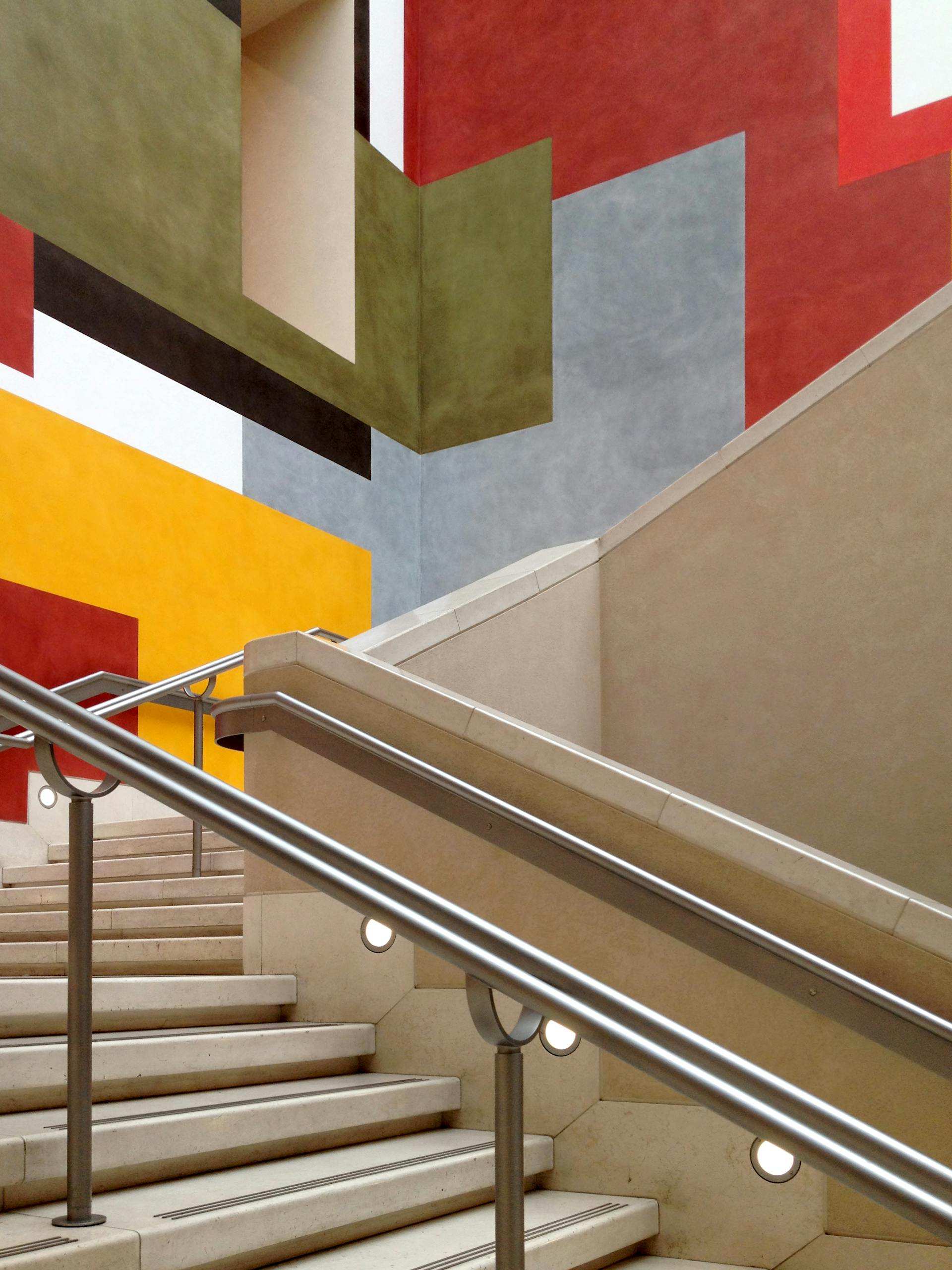 Escalier aux marches en pierre beige et à la rampe en acier épurée. Les murs environnants sont ornés de motifs géométriques audacieux en rouge, gris, vert et jaune, créant un décor vibrant et inspiré de l'art moderne. Un éclairage subtil accentue l'espace, mettant en valeur ses lignes architecturales.